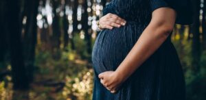 Mulher grávida vestindo um vestido azul escuro segura delicadamente sua barriga enquanto caminha em um ambiente natural com árvores ao fundo
