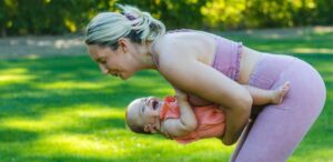 Mãe praticando atividade física ao ar livre enquanto segura seu bebê no colo, ambos sorrindo. Exercício no pós-parto melhora o bem-estar.