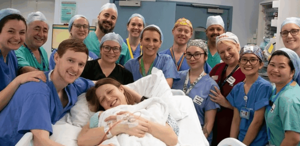 Equipe médica celebra nascimento após transplante de útero no Reino Unido, com mãe, pai e bebê saudáveis no centro da imagem.