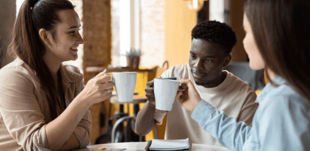Três pessoas conversam e brindam com canecas de café, representando o impacto da cafeína na interação social e no bem-estar mental.
