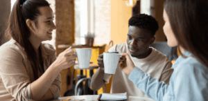 Três pessoas conversam e brindam com canecas de café, representando o impacto da cafeína na interação social e no bem-estar mental.