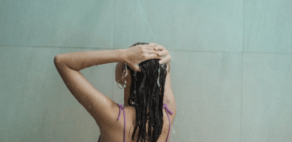 Mulher tomando banho, com água caindo sobre os cabelos, representando o impacto do banho na saúde da pele