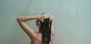 Mulher tomando banho, com água caindo sobre os cabelos, representando o impacto do banho na saúde da pele