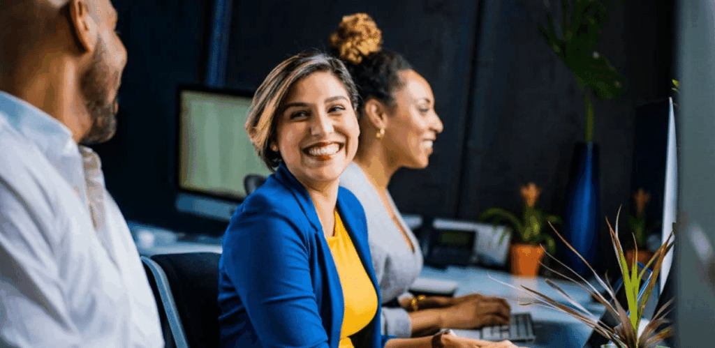 Três profissionais trabalhando em escritório moderno, sorrindo e demonstrando bem-estar durante o expediente