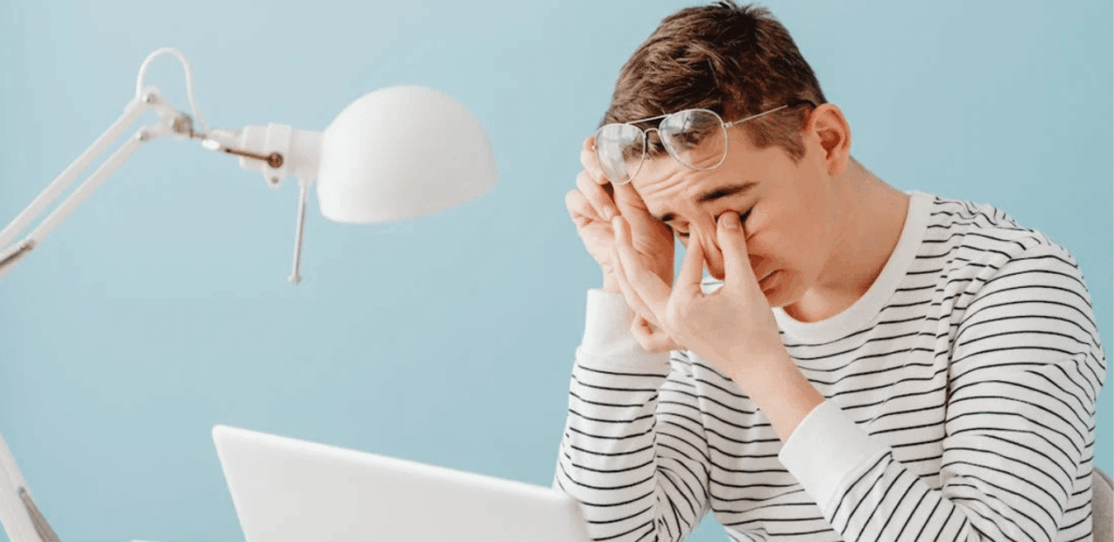 Jovem com sinais de fadiga ocular digital esfregando os olhos em frente ao computador, após longo tempo de tela.