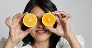 Mulher sorridente segurando laranjas como óculos, simbolizando alimentos que melhoram o humor